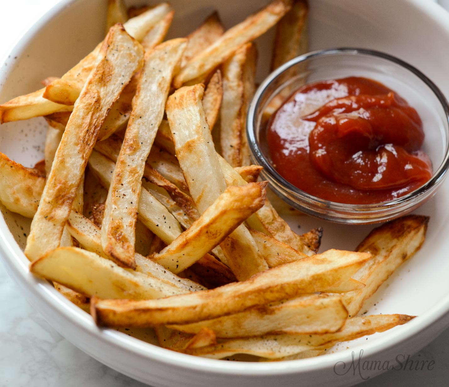 A white serving bowl filled with french fries made in an air fryer and a little serving bowl of ketchup.