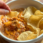 A tortilla chip being dipped into chili cheese dip.