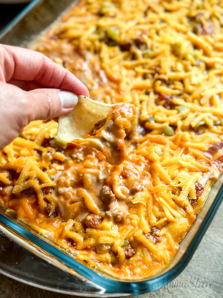 A glass casserole dish of baked chili cheese dip with a chip being dipped in. 