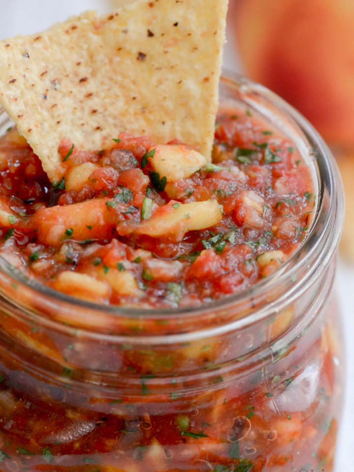 A closeup of a jar of homemade peach salsa with a tortilla chip dipped in. And the jar is slightly offset to the left in the picture.