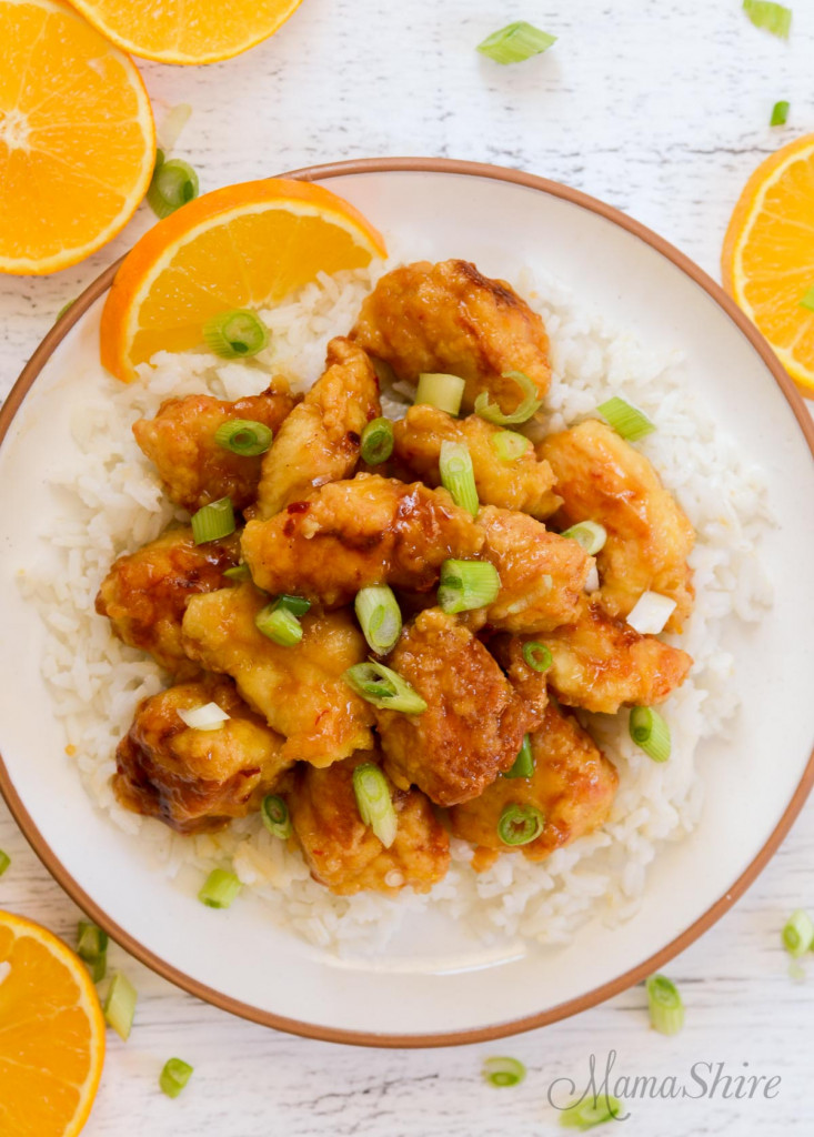 A plate of easy orange chicken on top on rice with some slices of orange and slices of green onions.