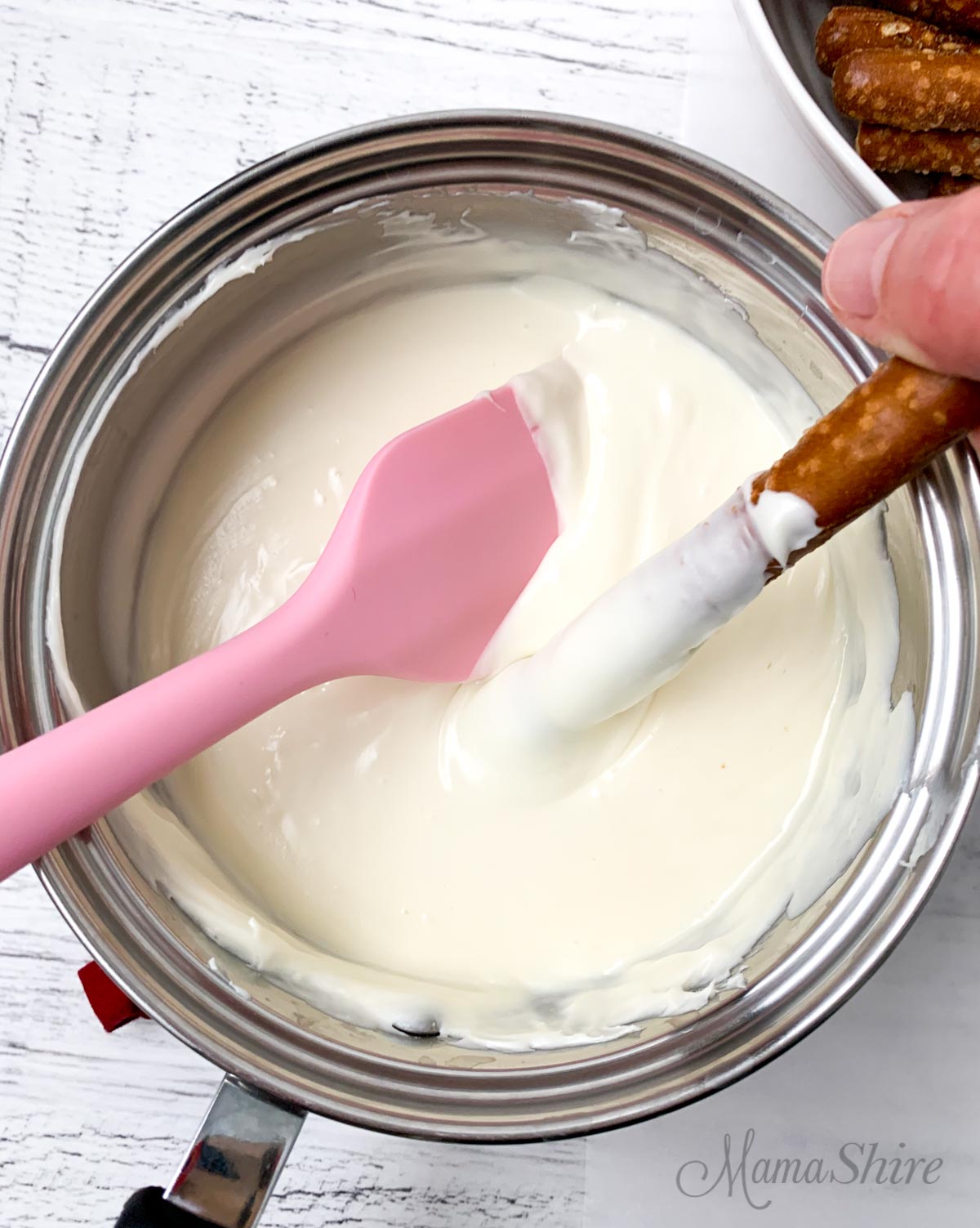 A pan with melted white icing and a pink spatula to help spread icing onto the pretzel rod.