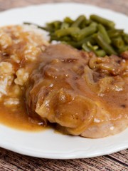 A vertical picture of a white dinner plate with a pork chop that has onion gravy poured over the top with a side of white rice and green beans.