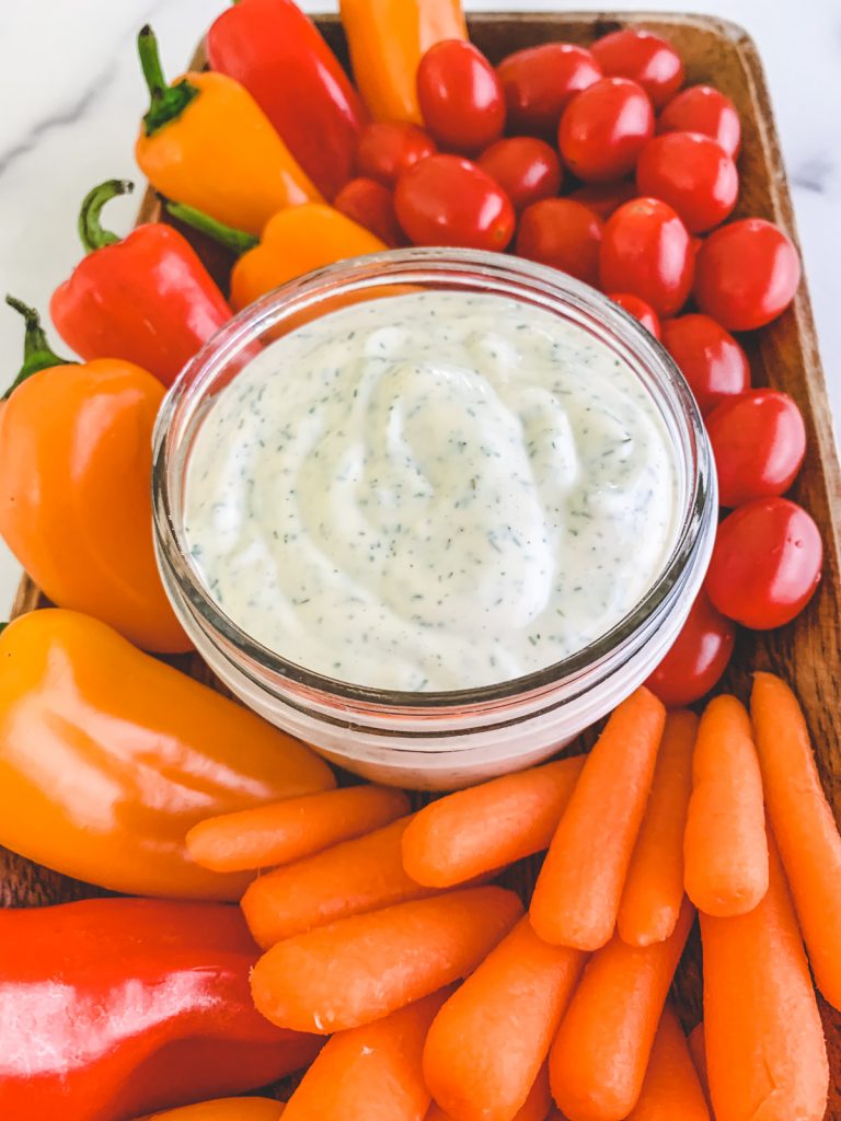 A serving of homemade dairy-free ranch dressing surrounded by veggies, like carrots, peppers, and cherry tomatoes.
