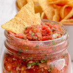 A ball jar with with restaurant style salsa with a tortilla chip sticking in the top.