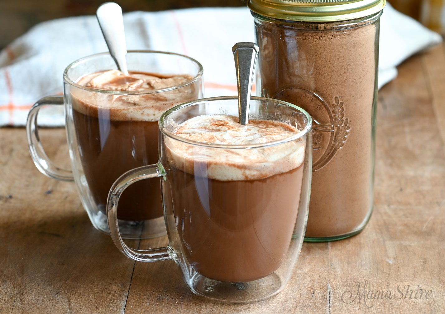 Two mugs of hot chocolate with a jar of dairy-free and gluten-free hot chocolate mix.