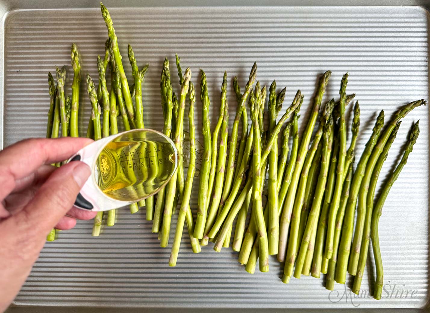 Oil being drizzled over asparagus on a baking pan.