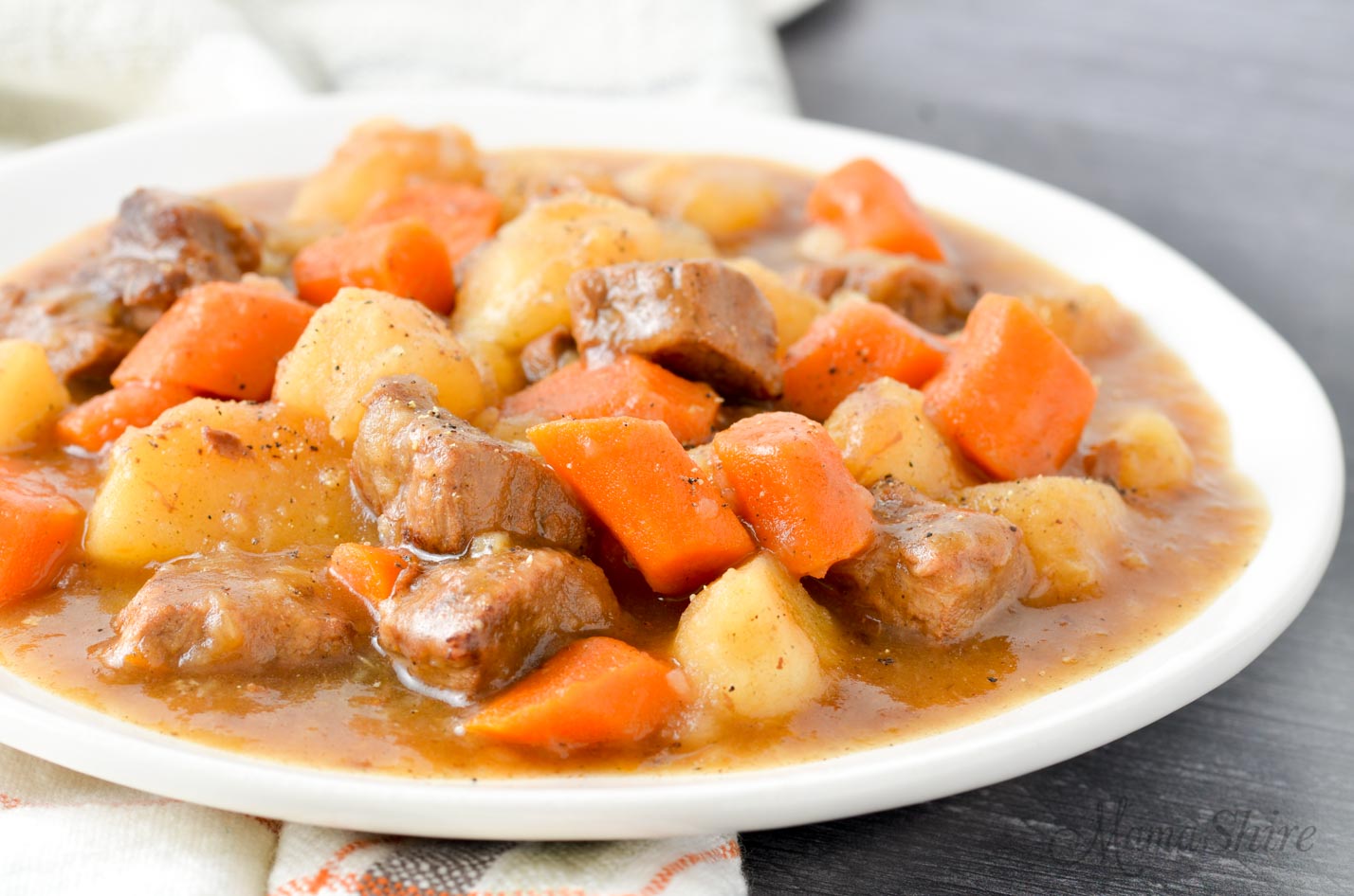 A serving of beef stew on a white plate that includes beef, carrots, potatoes, onions, and a nice thick gravy.