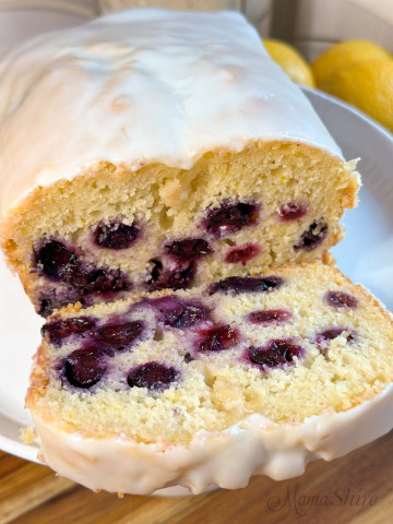 A loaf of sweet bread made with lemon juice and blueberries with a slice laying forward.