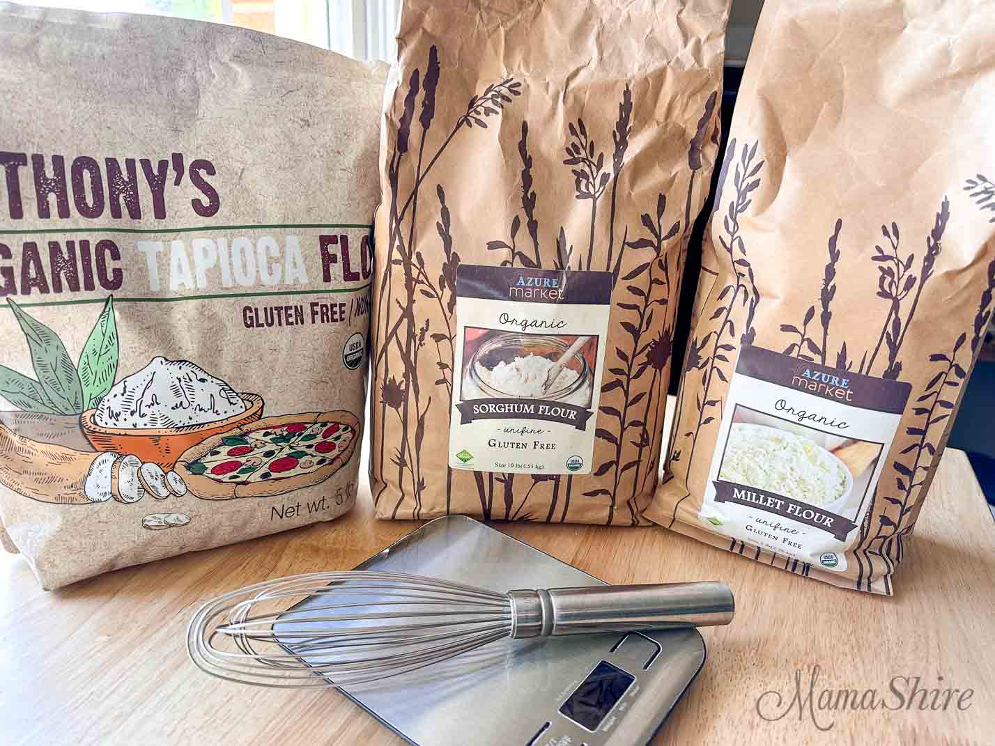Three bags of gluten-free flours. Tapioca, sorghum, and millet. With a food scale and wire whisk.