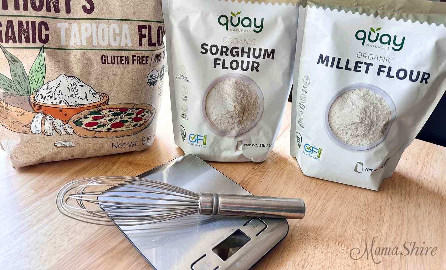 Three bags of flour, organic tapioca starch, organic sorghum, organic millet. And in front is a food scale and wire whisk. 