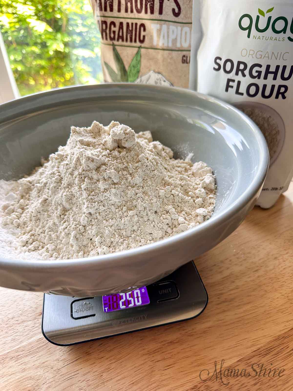 A large grey mixing bowl with organic tapioca starch and organic sorghum flour on top of a food scale. 