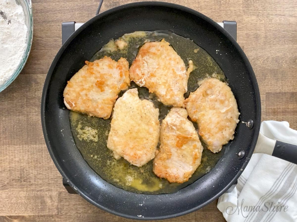 A skillet of thin pork chops frying that have a gluten-free flour coating.